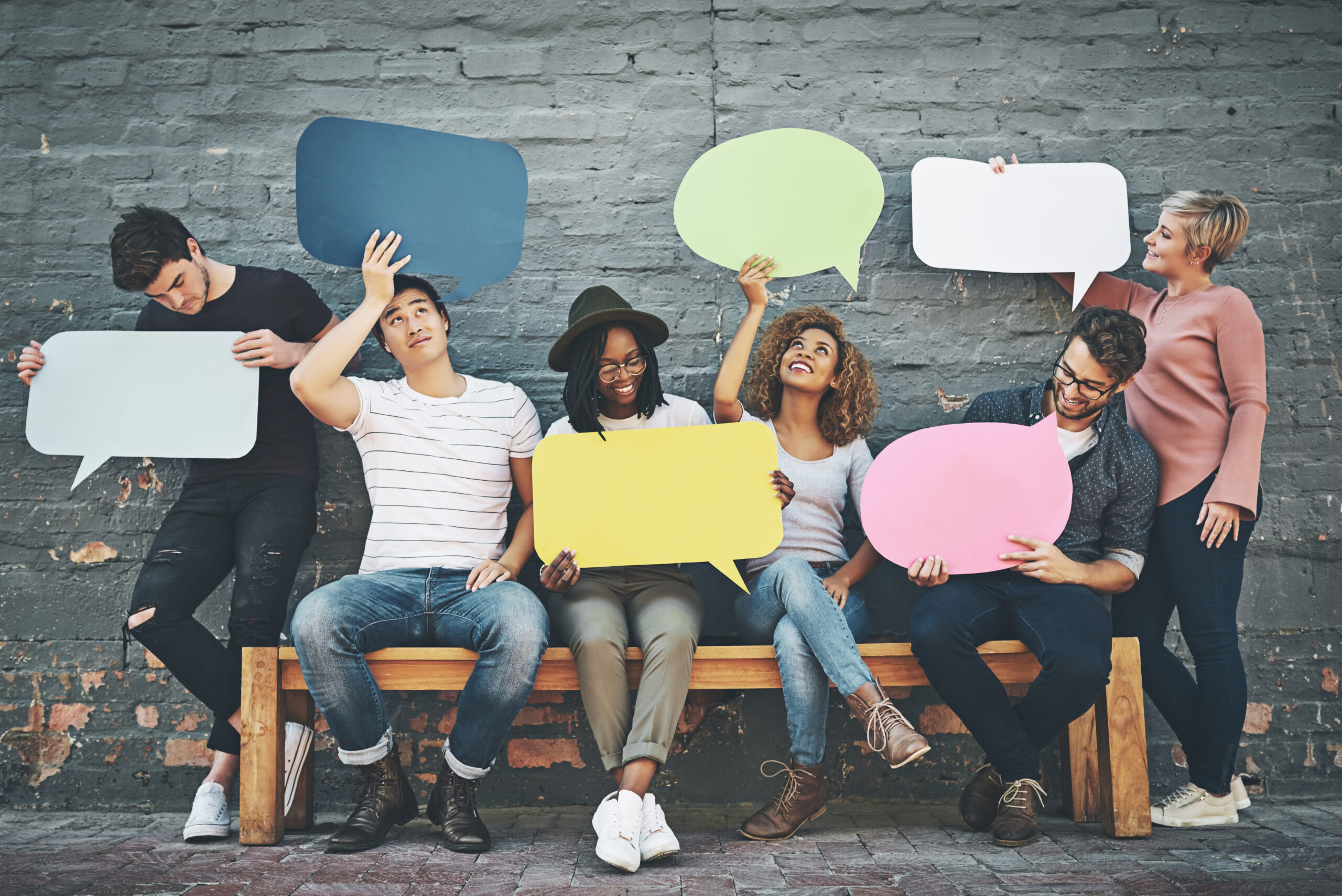 Diverse group of people sitting on a bench holding up blank speech bubbles.